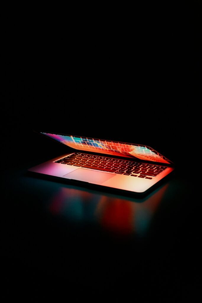pexels-photo-2047905-2047905 Laptop partially opened, emitting vibrant multicolored lights in dark setting.